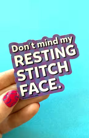 Purple plaid vinyl sticker reading “Don't mind my Resting Stitch Face,” held against a turquoise background.

